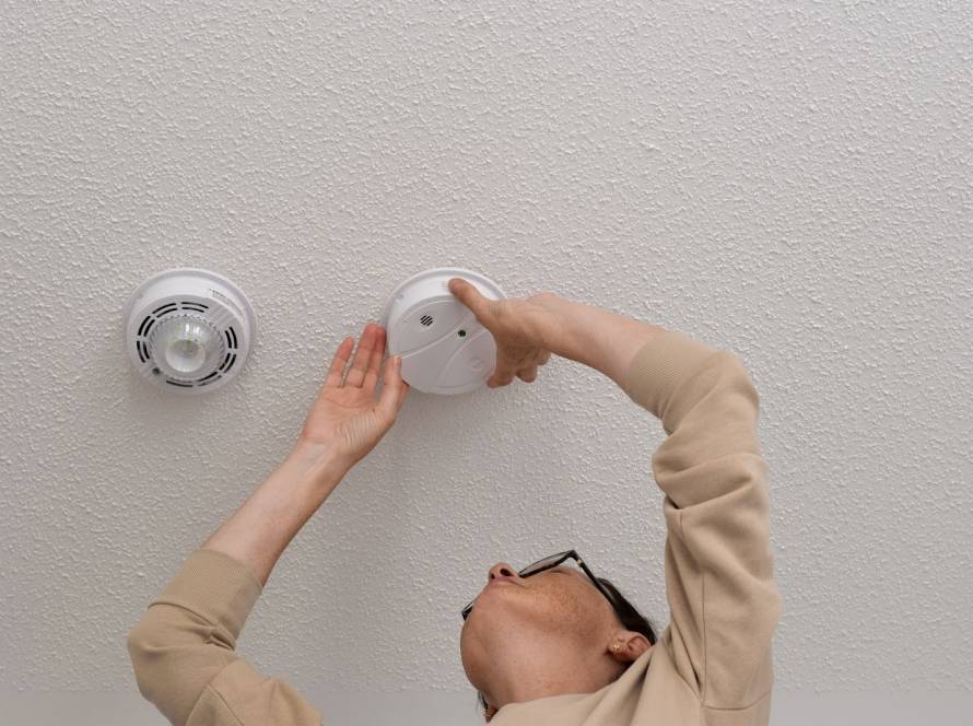 Alt-text: A technician’s hand mounting a carbon monoxide detector on a ceiling.