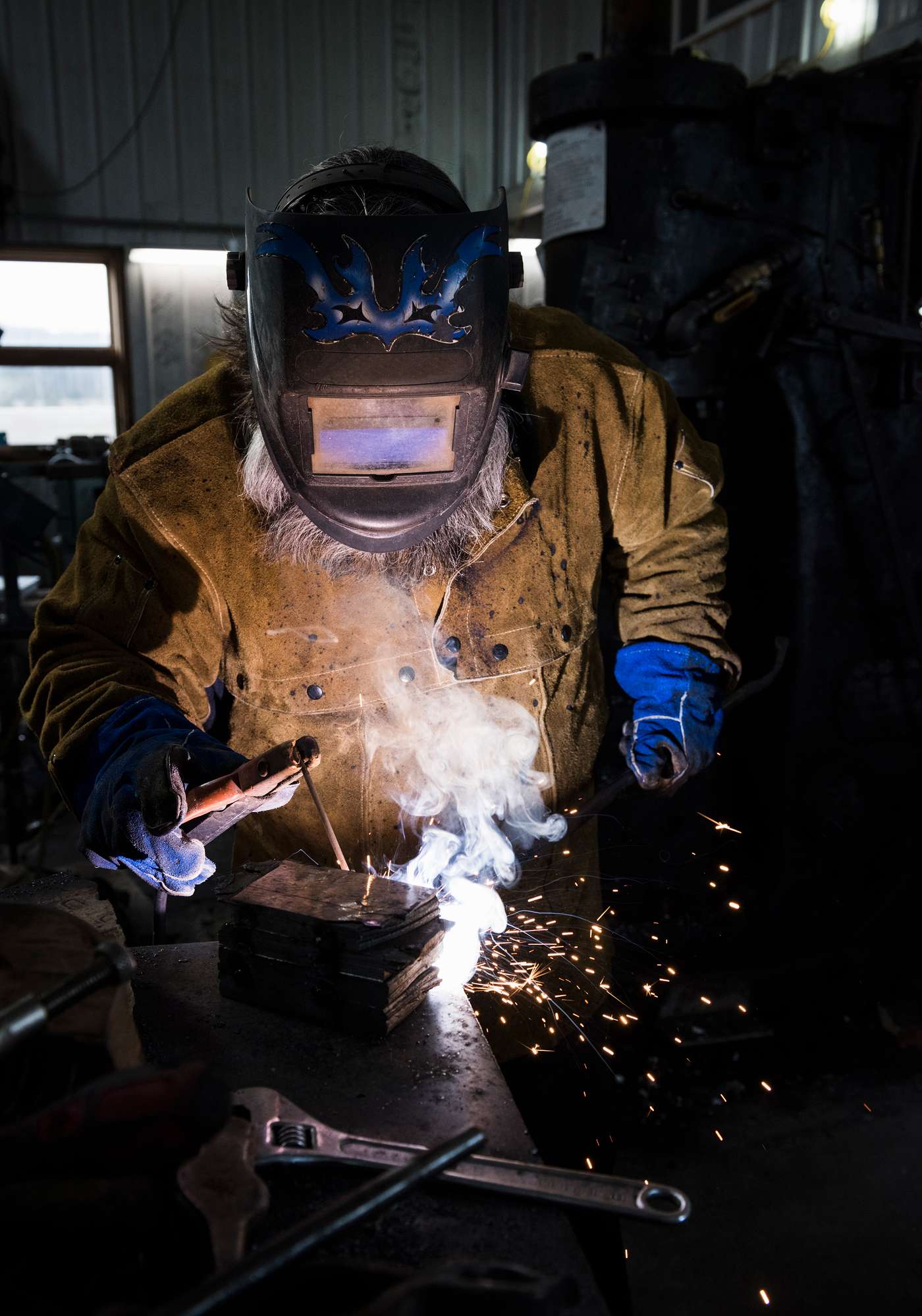 A welder in protective gear works on a metal piece, producing sparks and smoke in a workshop setting. The scene highlights the skill and precision involved in welding.