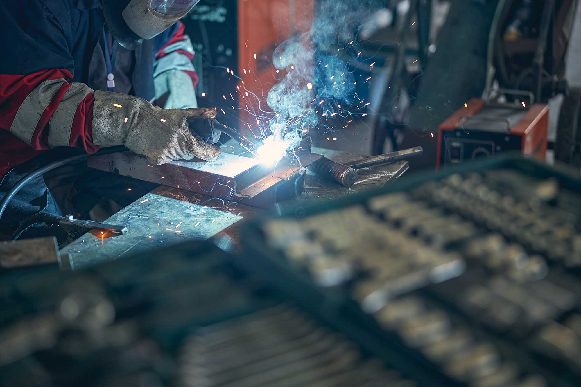 A welder in protective gear works on a metal piece, creating sparks and smoke during the welding process. The scene captures the tools and equipment used for metal repairs in a workshop setting.