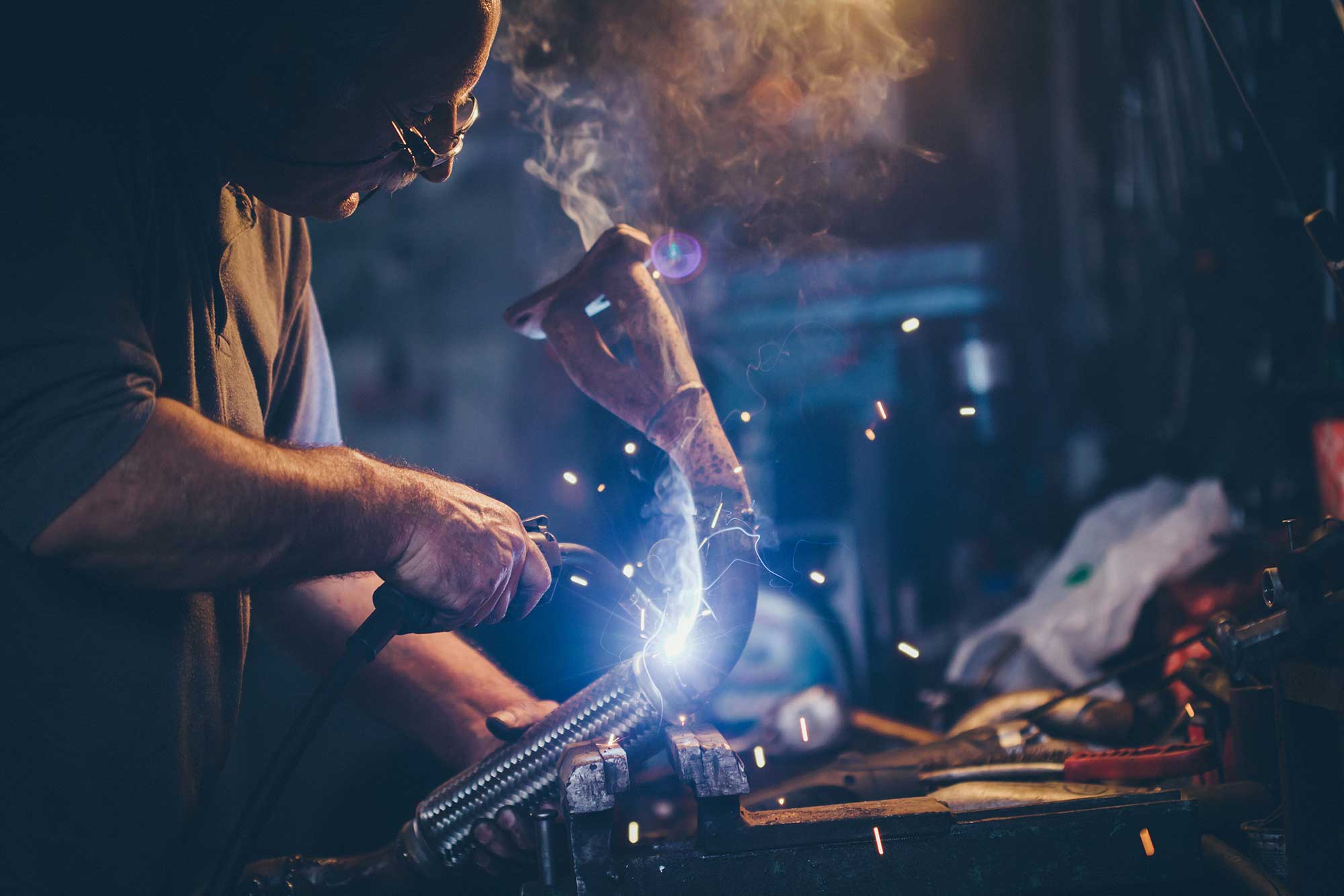 A welder repairs a metal part, creating sparks and smoke in a dimly lit workshop. The focus is on the welding process, demonstrating the skill and precision involved in metal repair.
