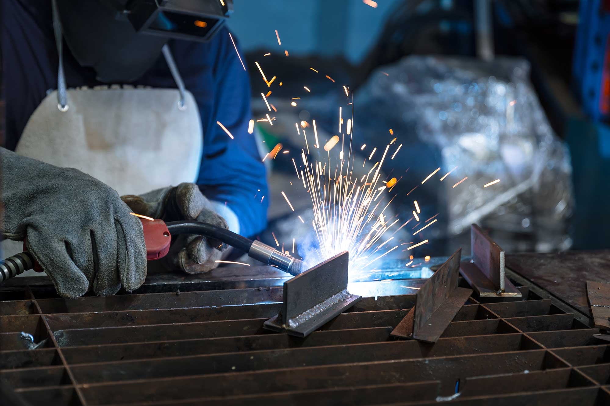 A welder uses a welding torch to join metal pieces, creating sparks in a workshop setting. The focus is on the precision and care involved in steel fabrication.