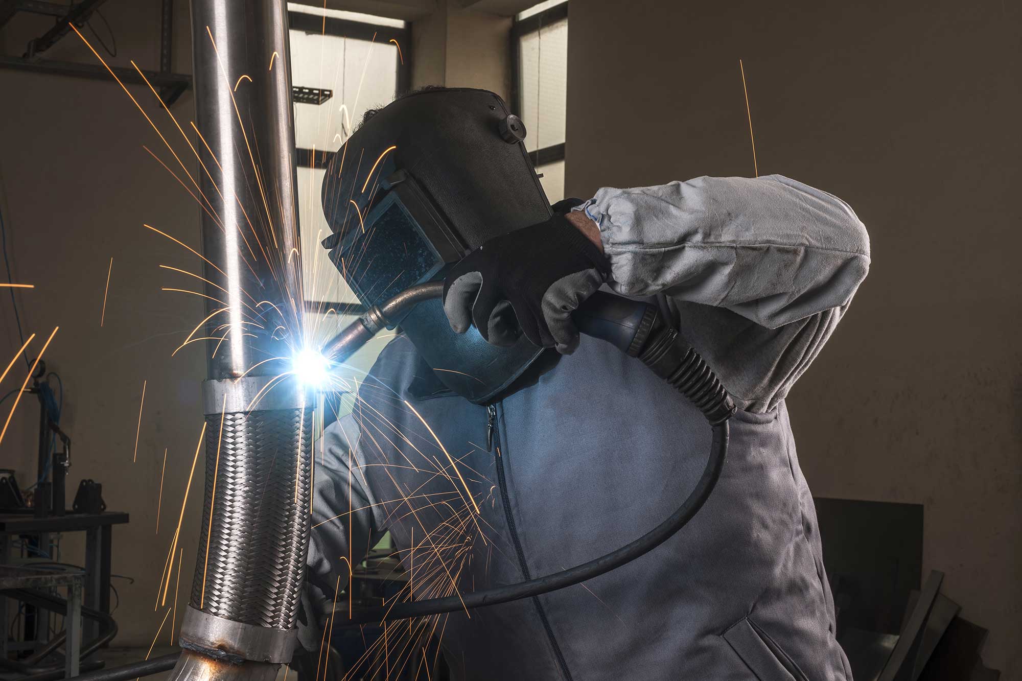 A welder wearing protective gear uses a welding torch to join aluminum pieces in a workshop, with sparks flying from the weld. The environment is industrial, highlighting the specialized process of aluminum welding.