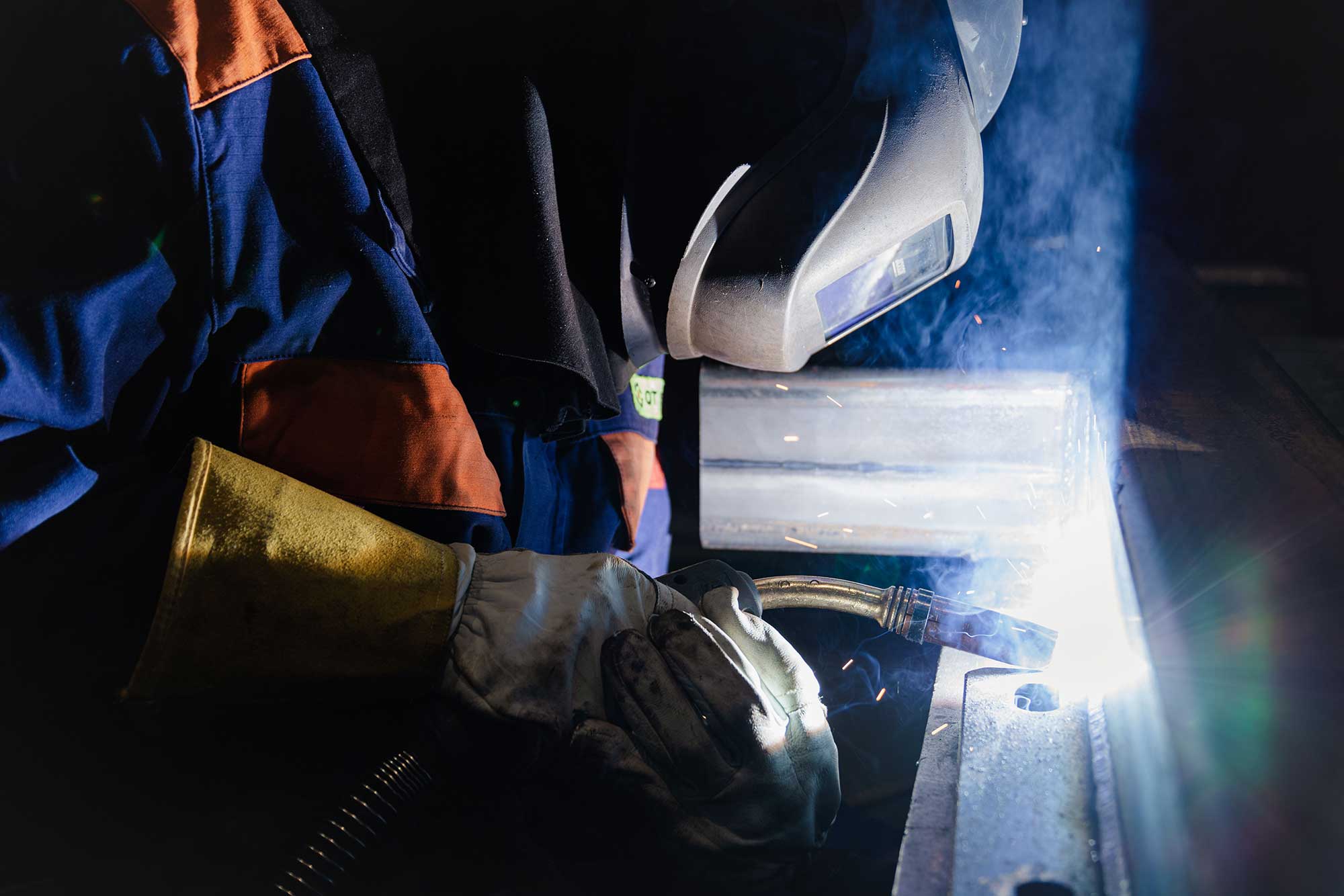 A welder in protective gear works on a metal piece, with sparks flying and smoke rising from the welding process. The setting appears to be dimly lit, highlighting the welding activity.
