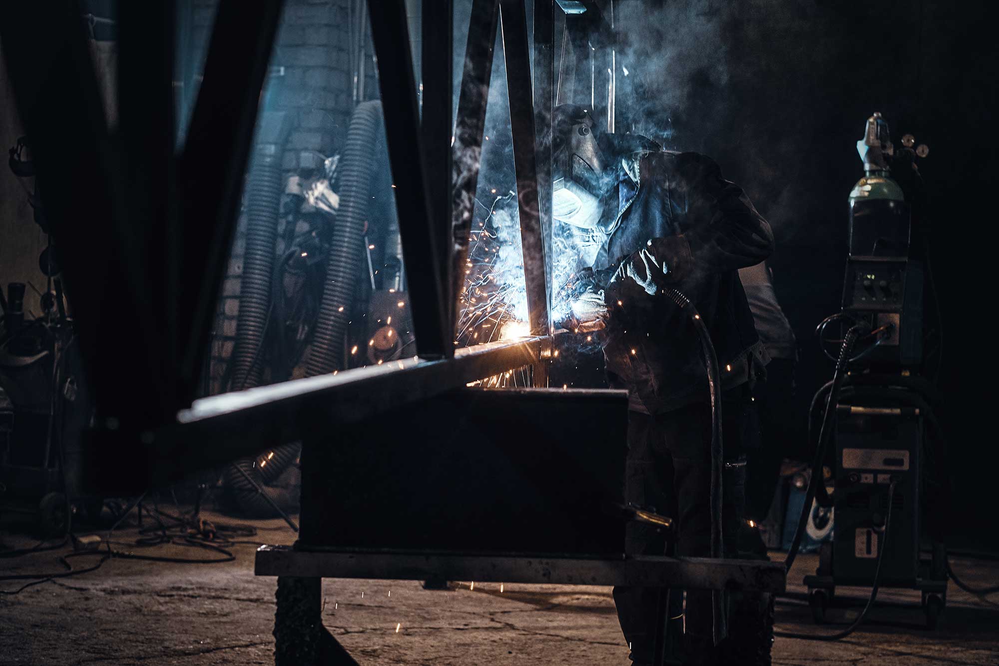 A welder works on a steel fabrication project in a workshop, with sparks flying and smoke in the air. The scene highlights the process of shaping and welding metal components.
