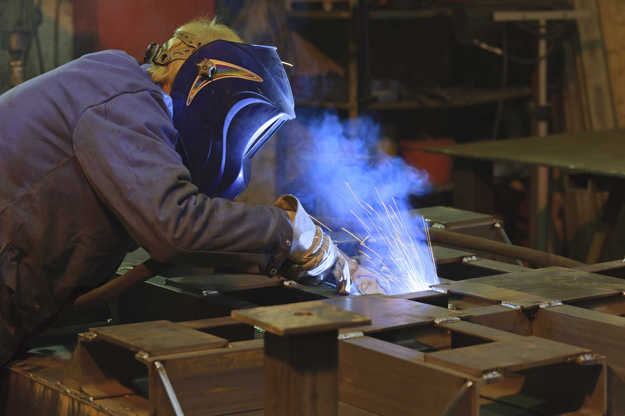 A welder in protective gear works on a metal structure, creating strong joints with sparks and smoke visible. The scene highlights the process of steel fabrication in a workshop setting.
