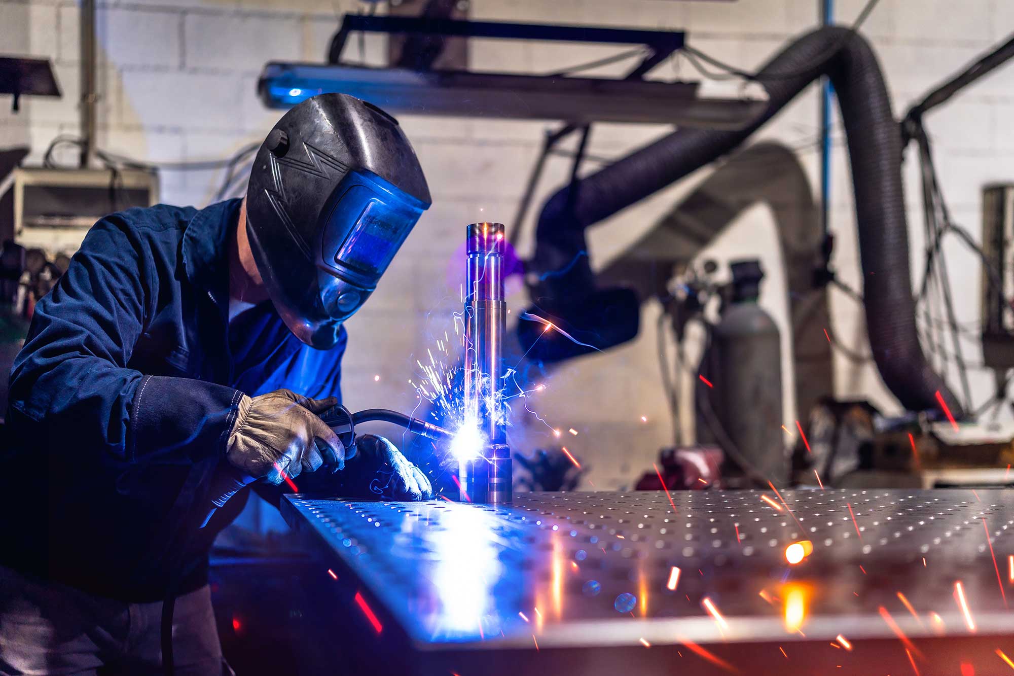 A welder wearing a protective helmet works on a piece of aluminum, producing bright sparks as they create a strong weld. The setting appears to be a workshop with various welding equipment in the background.