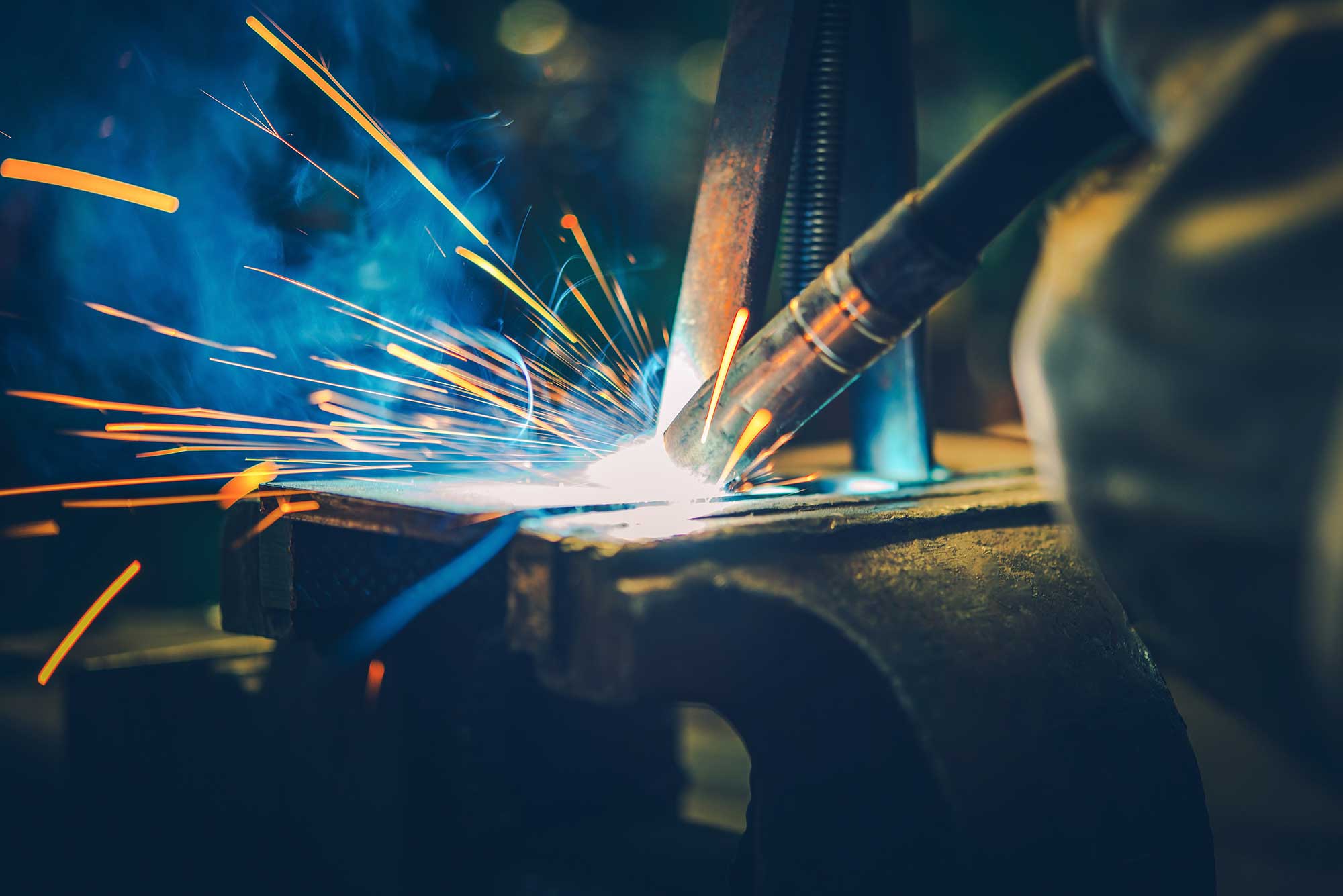 A welder uses a welding torch, creating bright sparks and blue flames as they work on a metal piece. The scene captures the intensity and precision of steel fabrication.