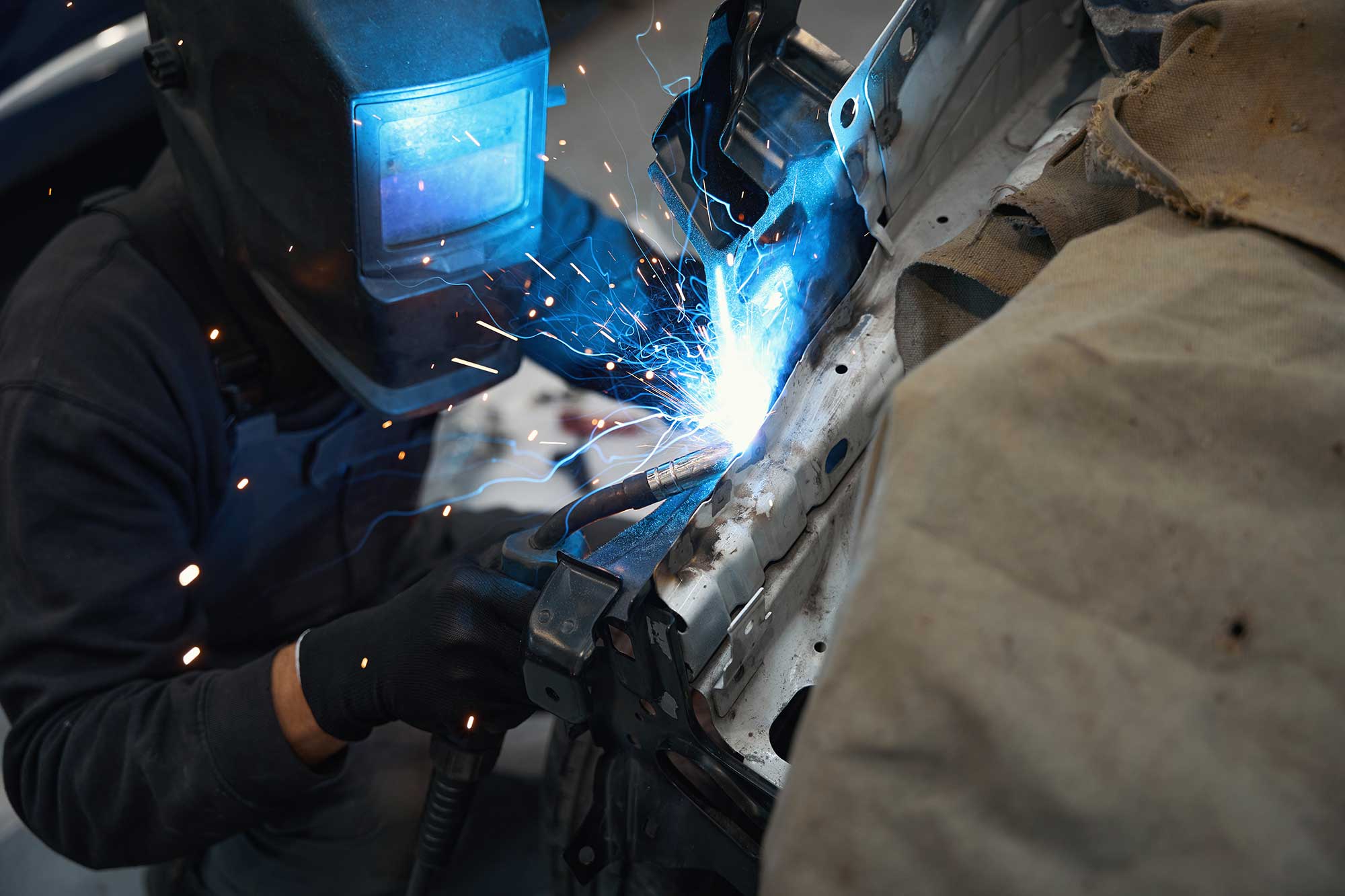 A welder wearing a protective helmet works on repairing metal parts, with bright sparks flying from the welding process. The scene showcases the precision and skill involved in metal repair.