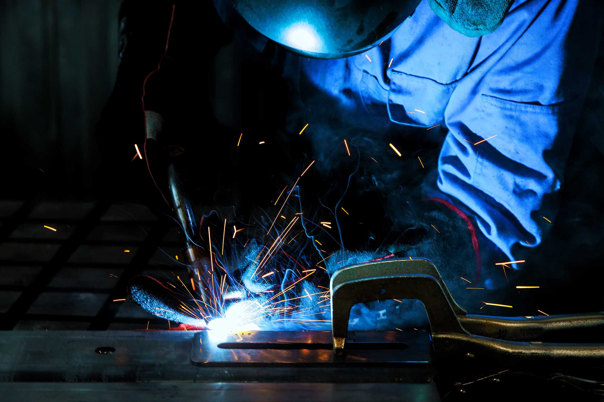 A welder in protective gear works on a metal piece, creating sparks and bright light from the welding process. The scene captures the precision and intensity of mobile welding services.