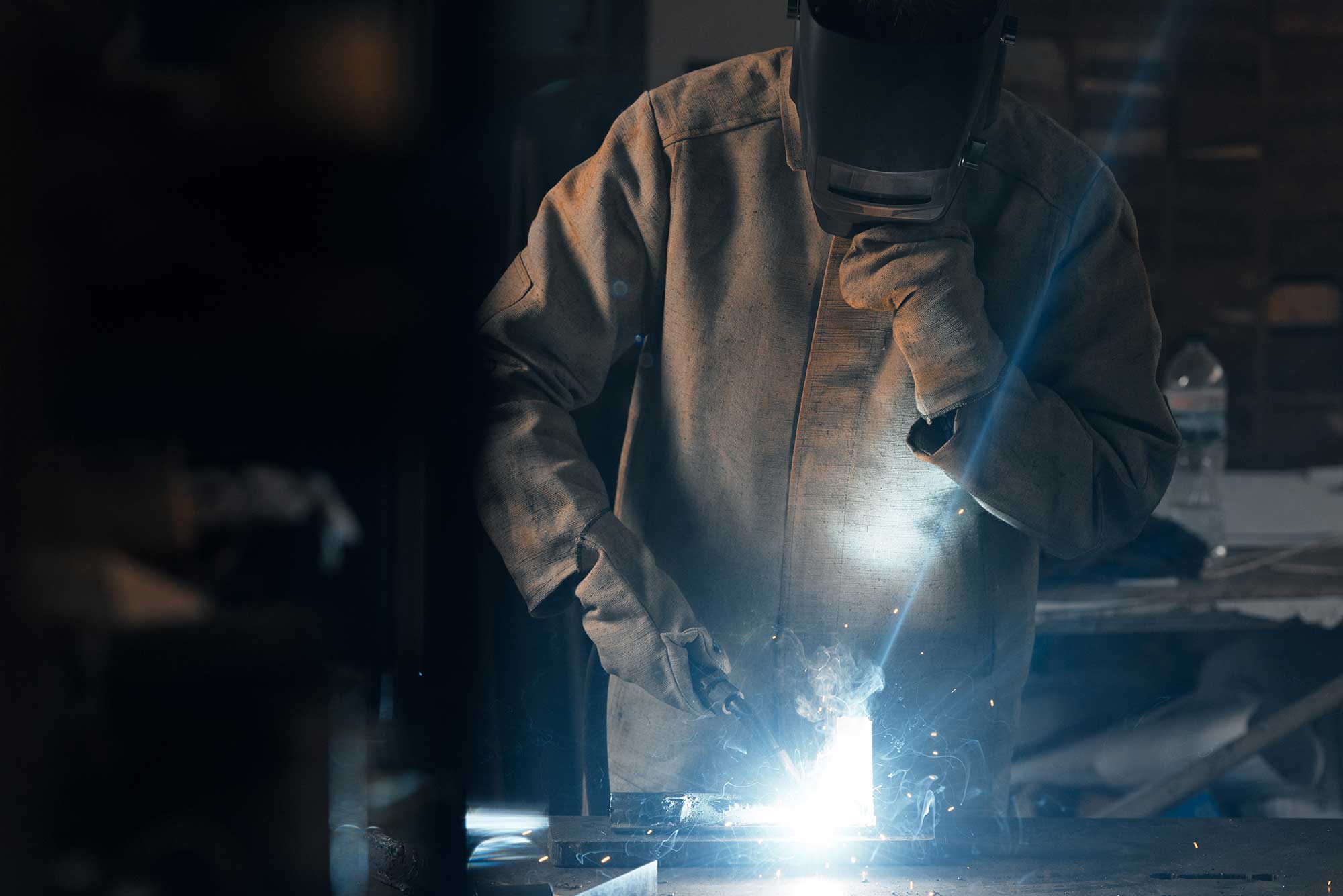 A welder in protective gear works on a metal piece, creating sparks and bright light as they perform emergency welding repairs. The scene highlights the urgency and skill involved in restoring metal structures.
