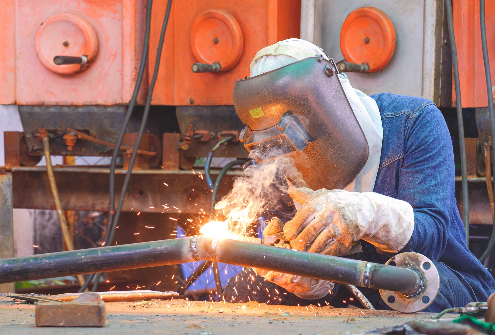 A welder wearing a protective mask works on a metal pipe, creating sparks and smoke as they perform emergency welding repairs in a workshop setting.