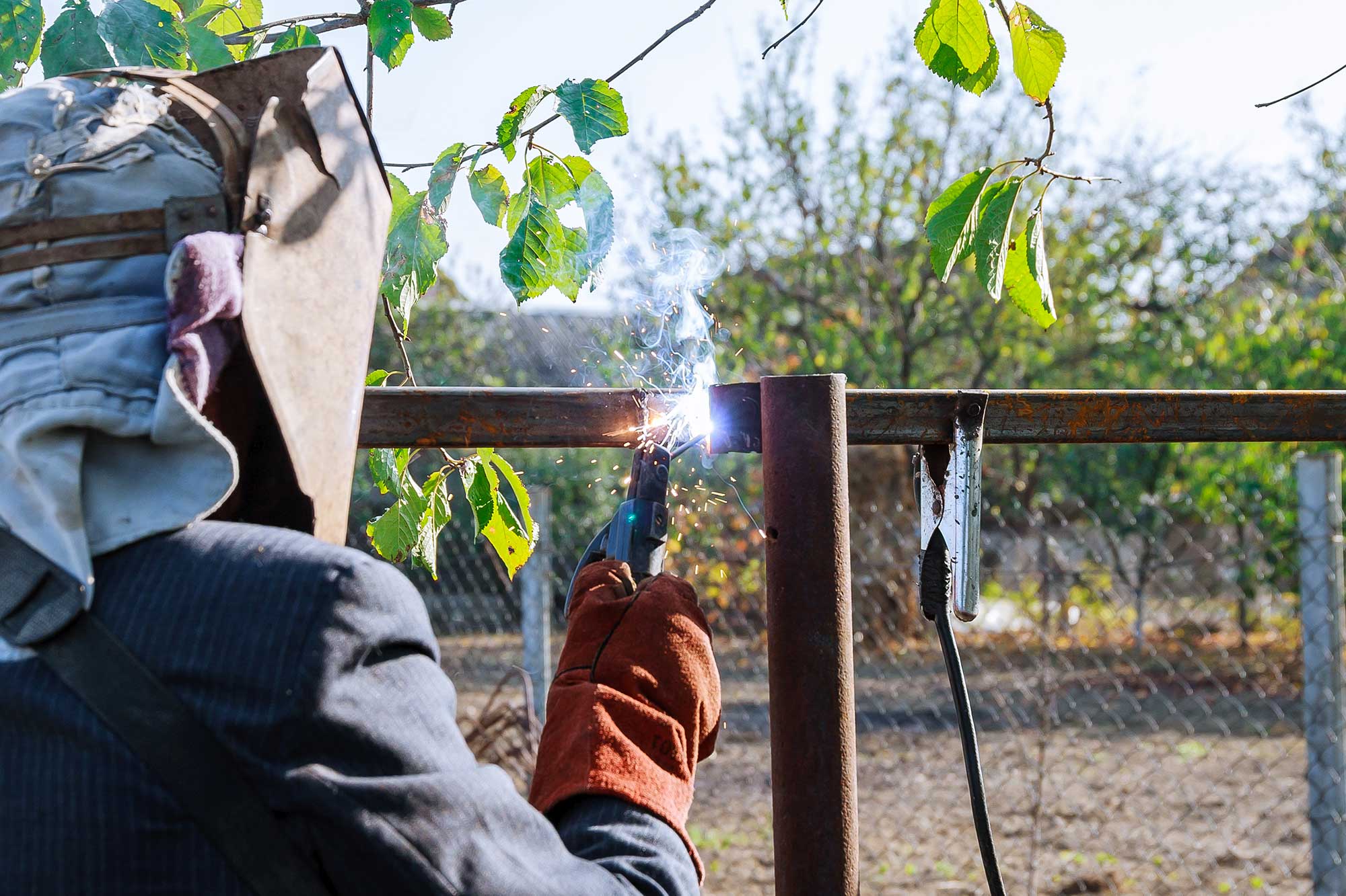 A welder in protective gear is working on a metal fence, using a welding torch that emits a bright spark. The background shows trees and a clear sky, indicating an outdoor setting.