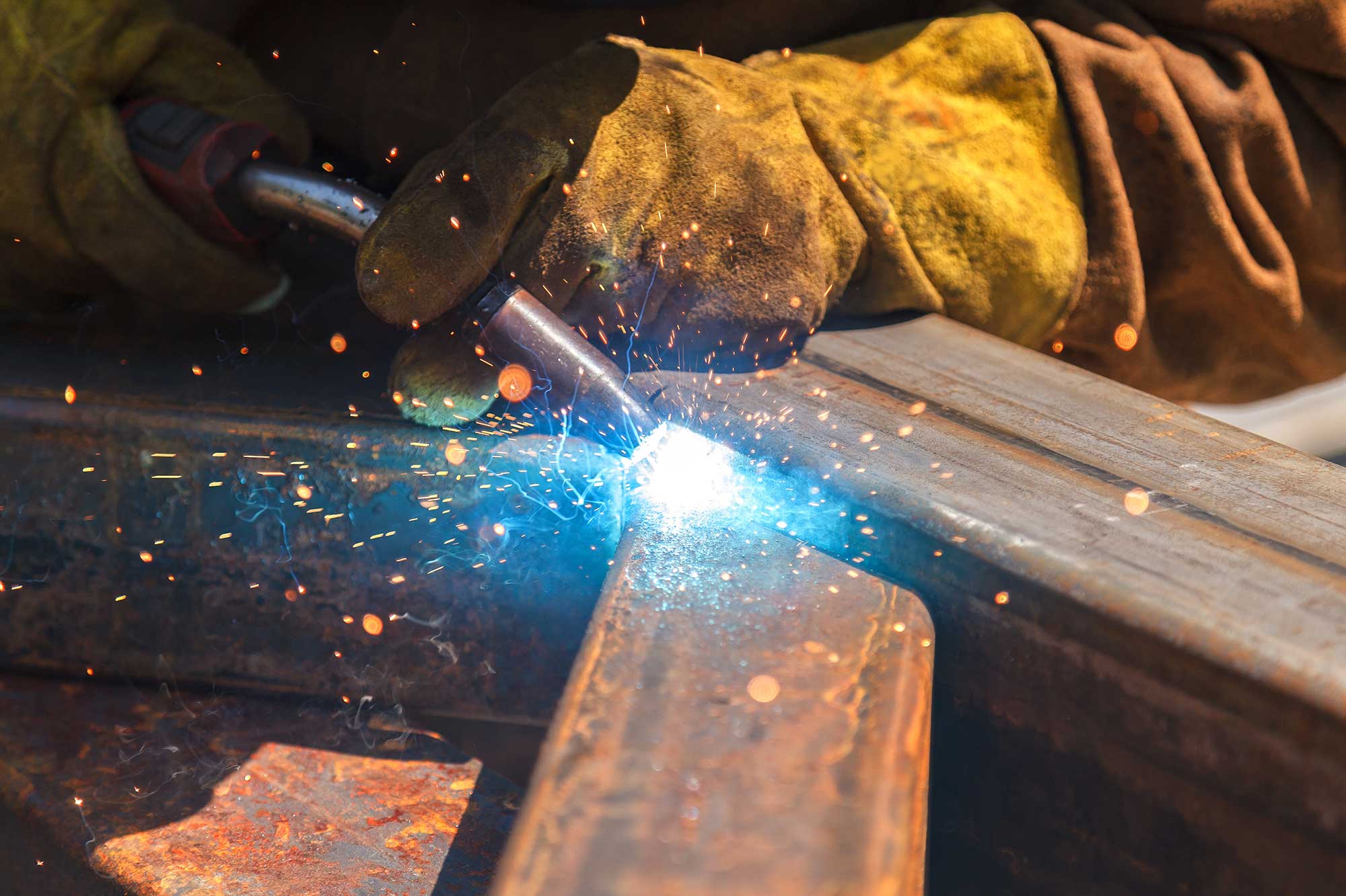 A welder uses a torch to join two metal pieces, creating bright sparks and a blue flame. The welder is wearing protective gloves and working on-site.
