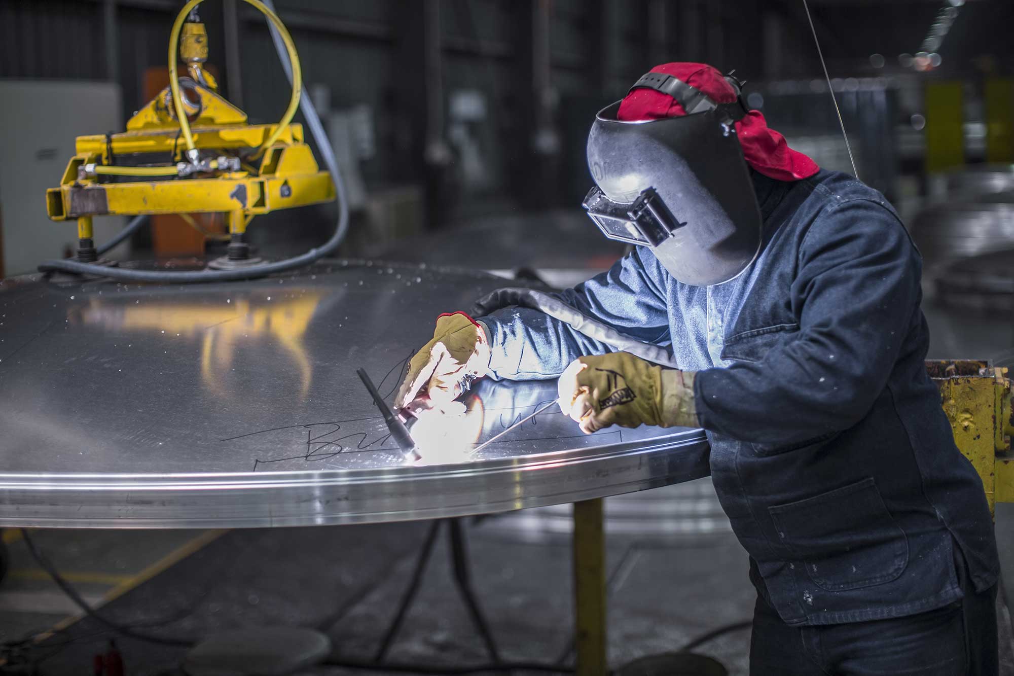 A welder in protective gear works on a large aluminum piece in a workshop, using a welding tool to create a strong joint. The environment is well-lit, showcasing the precision required for aluminum welding.