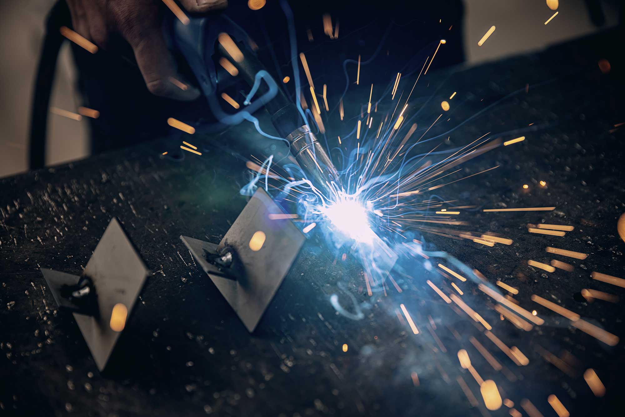 A welder uses a torch to create sparks while repairing metal parts on a work surface. The image captures the intensity of the welding process, highlighting the bright arc and flying sparks.